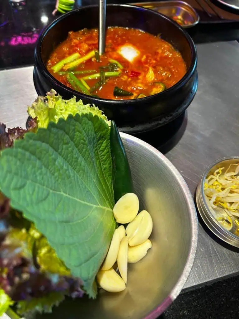 Primer plano de un soondubu jjigae (sopa coreana de tofu suave) hirviendo en olla de piedra negra, acompañado de un bowl con hojas de perilla, lechuga y dientes de ajo crudos para armar ssam, y brotes de soja al fondo.