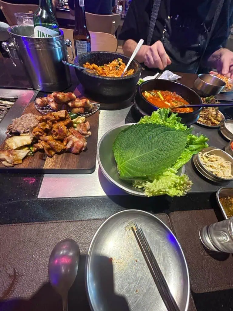 Vista general de la mesa de un restaurante de Korean BBQ: tabla de madera con carnes mixtas asadas, olla de piedra con arroz frito (bokkeum-bap) que una persona revuelve al fondo, bowl con hoja de perilla grande, sopa picante roja, palillos y banchan.