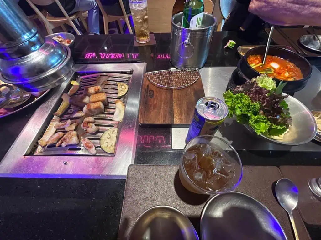 Vista cenital de una parrilla de Korean BBQ con samgyeopsal (panceta de cerdo) y otras carnes en distintas etapas de cocción, rodeada de vasos con bebidas, ensalada de hojas verdes y acompañantes en bowls de metal sobre una mesa oscura.