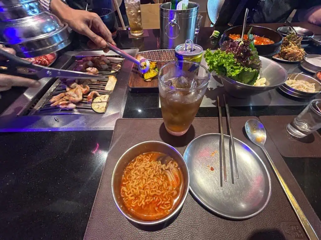 Tazón de ramyeon coreano en caldo rojo picante sobre la mesa, con palillos apoyados en un plato lateral; al fondo, una mano sostiene pinzas sobre la parrilla con carnes asadas, bebidas y hoja de perilla en un bowl de metal.